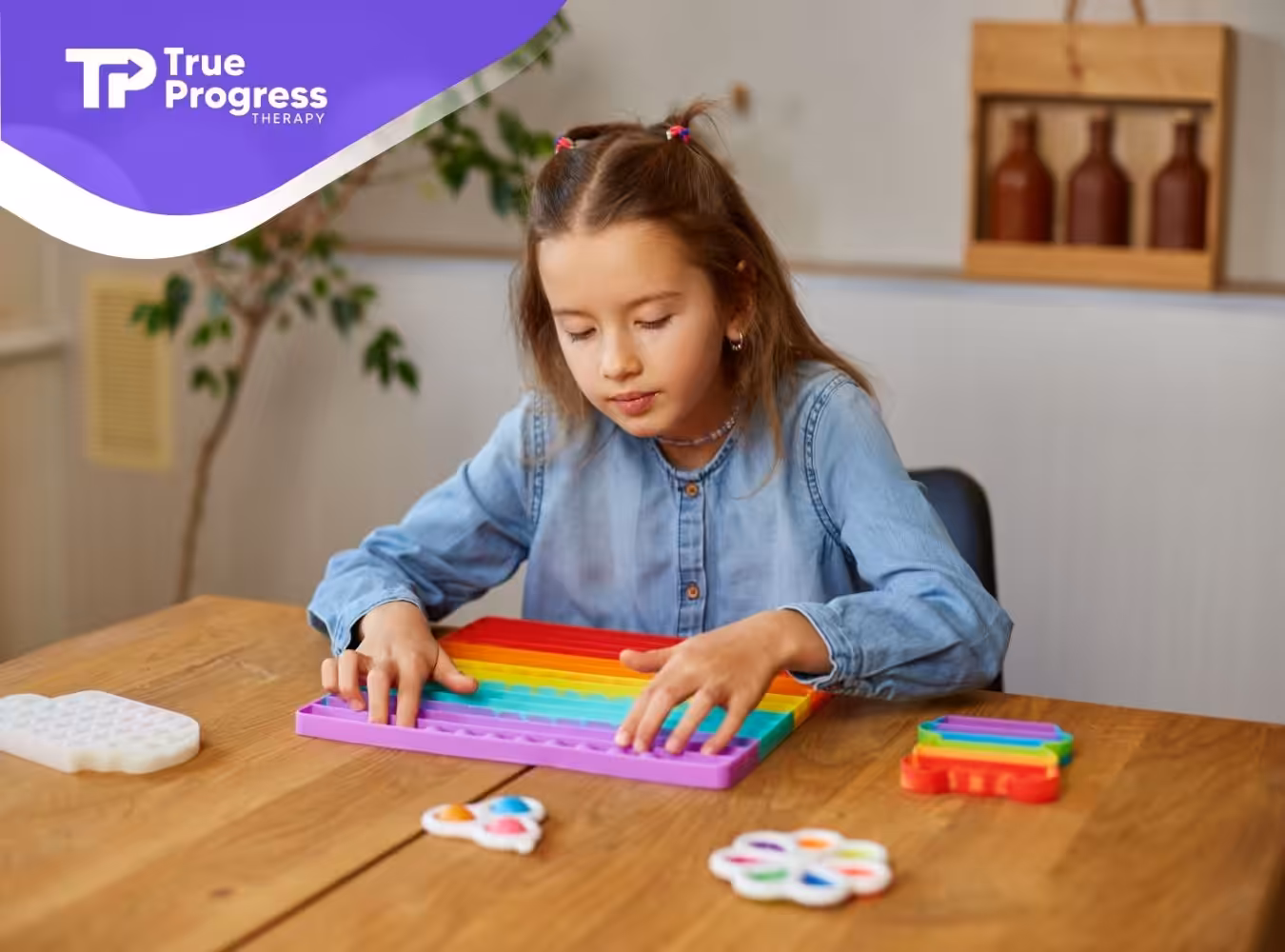 Young girl in a denim shirt focused on playing with a rainbow-colored pop-it at a wooden table.