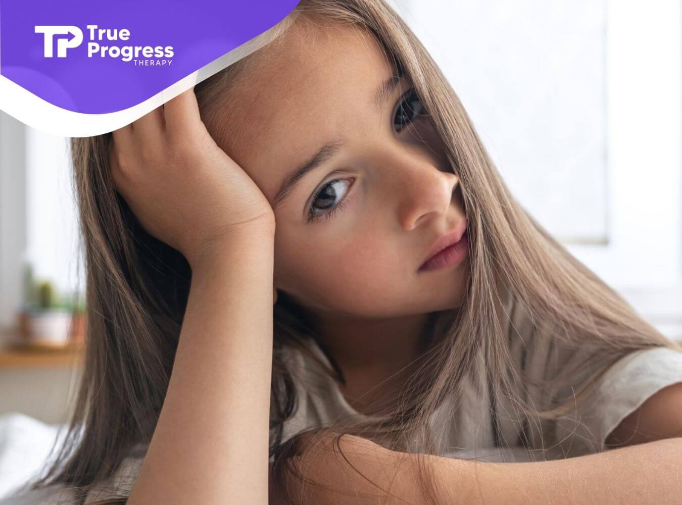 A young autistic girl with long brown hair rests her head on her hand.