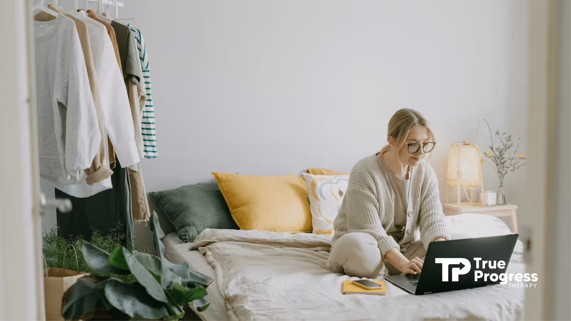 A BCBA sitting on a bed using a laptop in a softly lit, cozy bedroom, working remotely at home in NJ.
