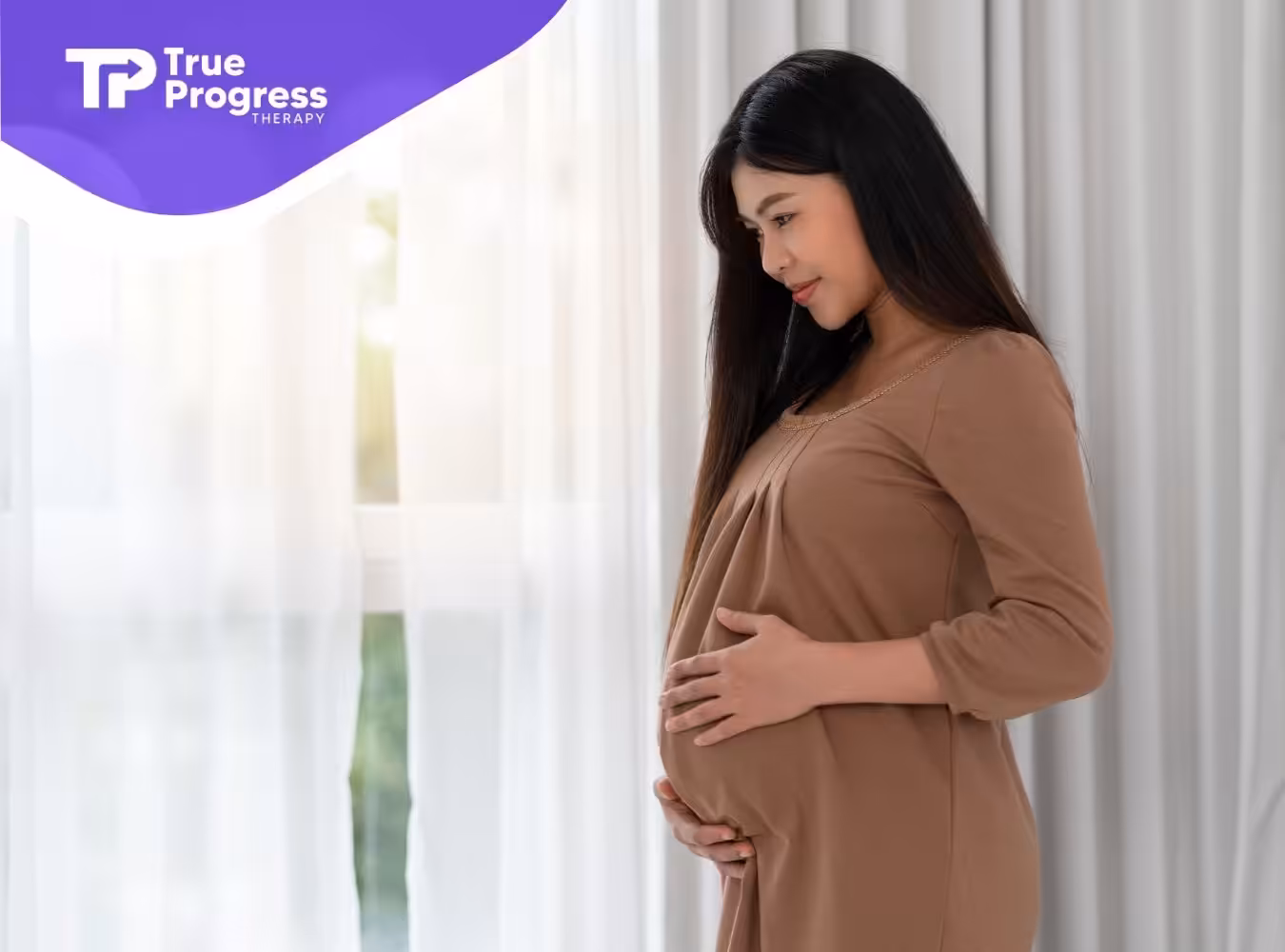 A pregnant woman in New Jersey gently holding her belly while standing in front of a bright window.