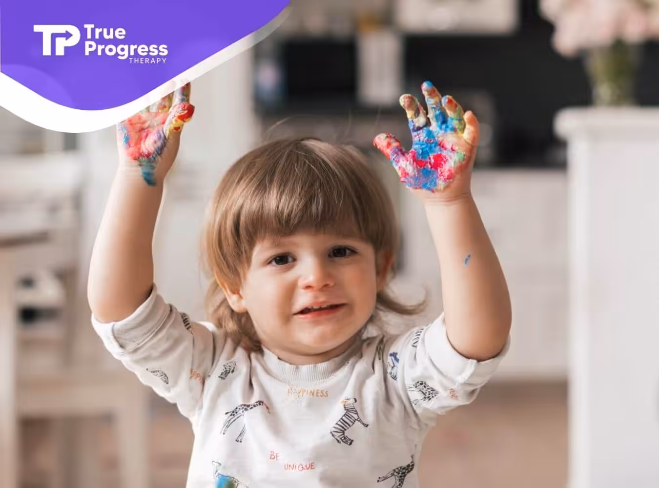 A toddler with autism with colorful paint on their hands excitedly raises them in the air.