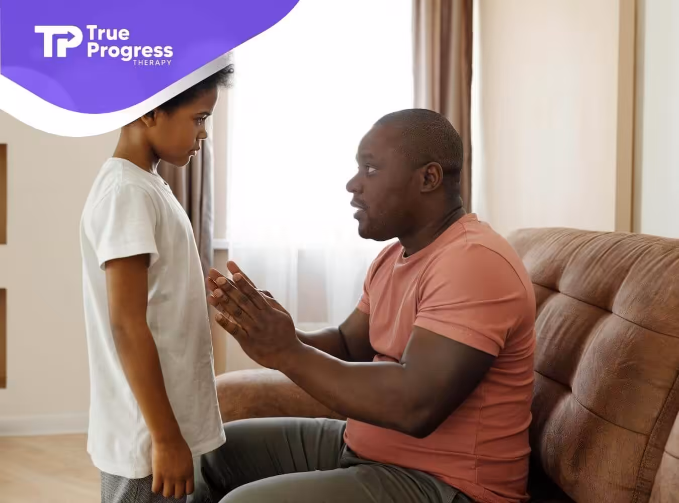 Man sits on couch, gesturing as he speaks seriously to child standing nearby in a softly lit home setting.