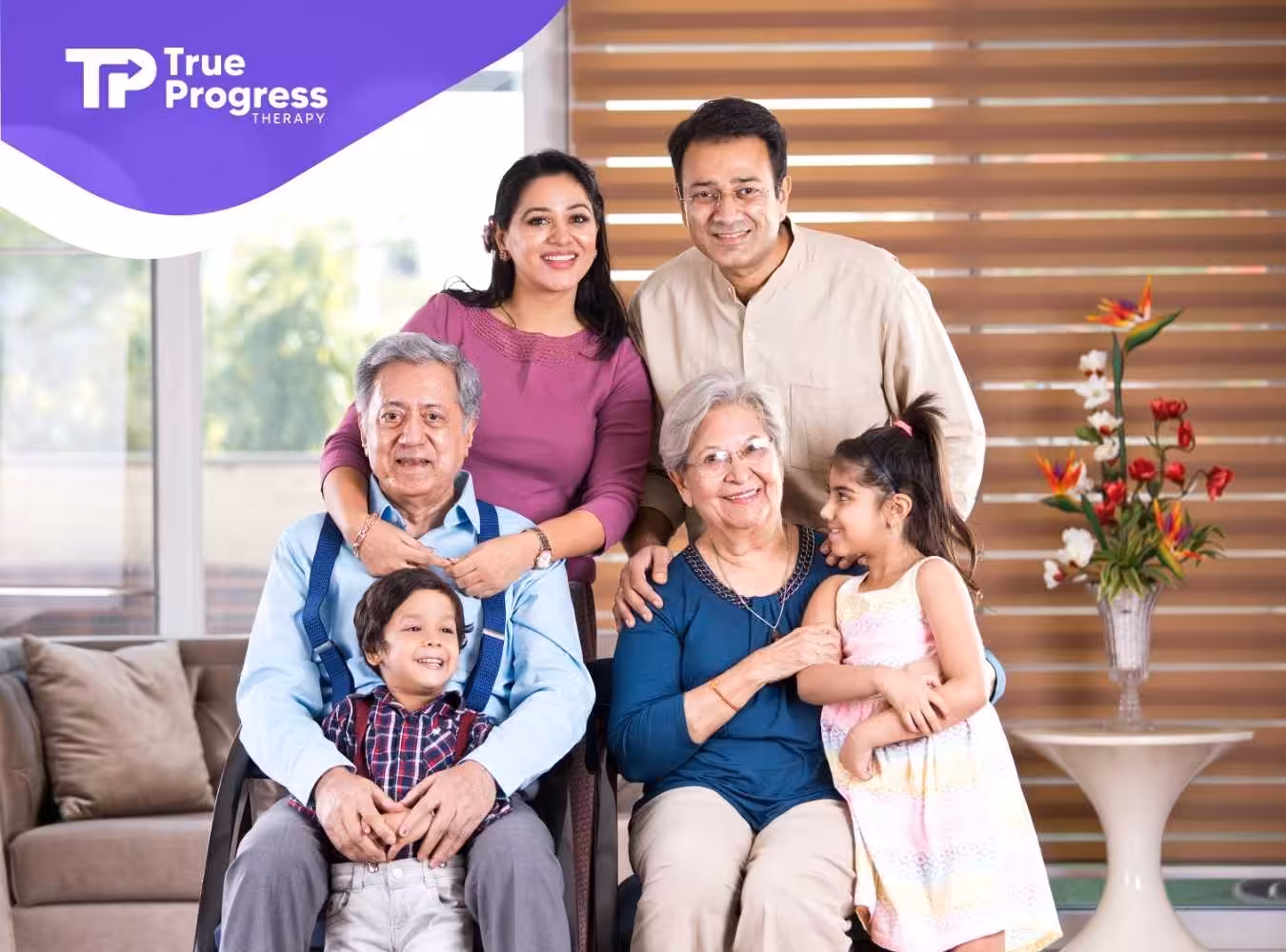 A multigenerational family of six smiling indoors. Grandparents are seated with two children beside them, while parents stand behind, exuding warmth and happiness.