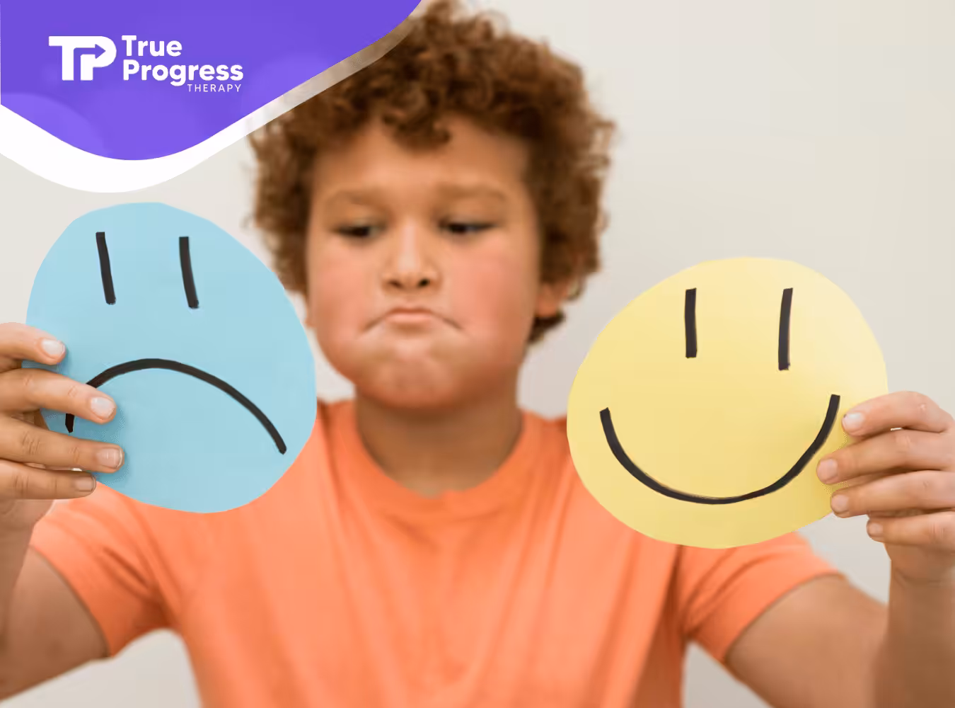 A child holding a blue sad face and a yellow happy face, appearing thoughtful. Wears an orange shirt, set against a neutral background.