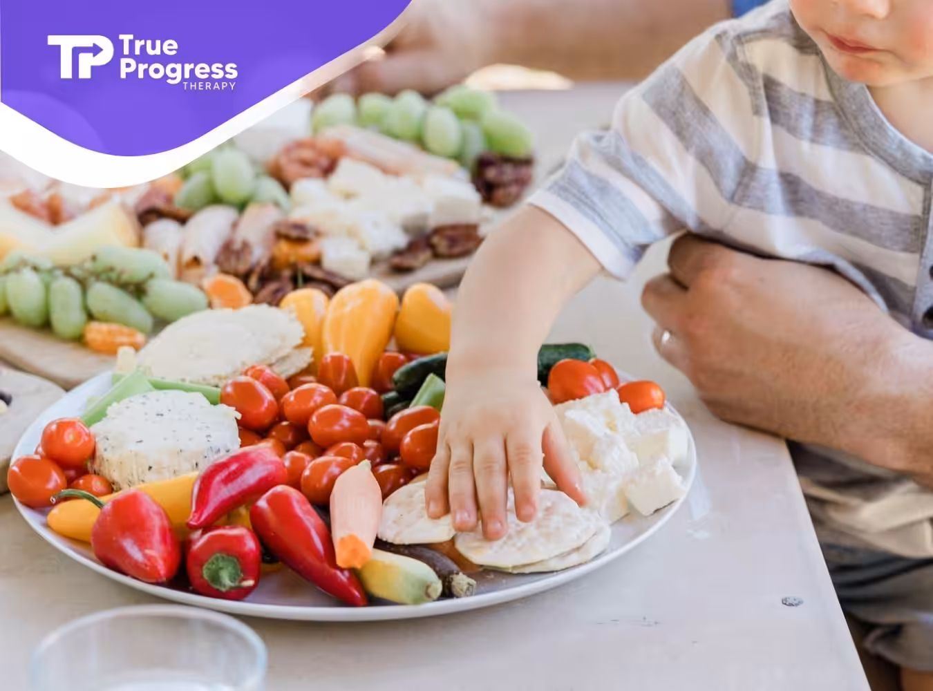 A child’s hand reaches for vegetables and cheese on a colorful platter, evoking a warm, family picnic setting.