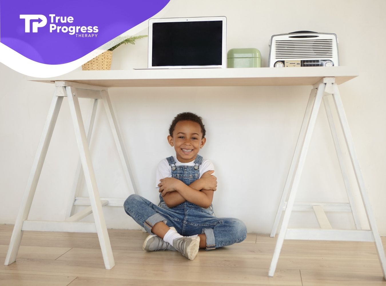 Smiling child sitting on the floor with arms crossed under a desk.