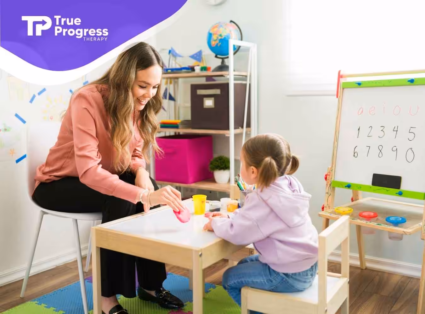 A smiling teacher interacts with a young girl at a small table in a colorful classroom. Behind them, an easel displays numbers and vowels.