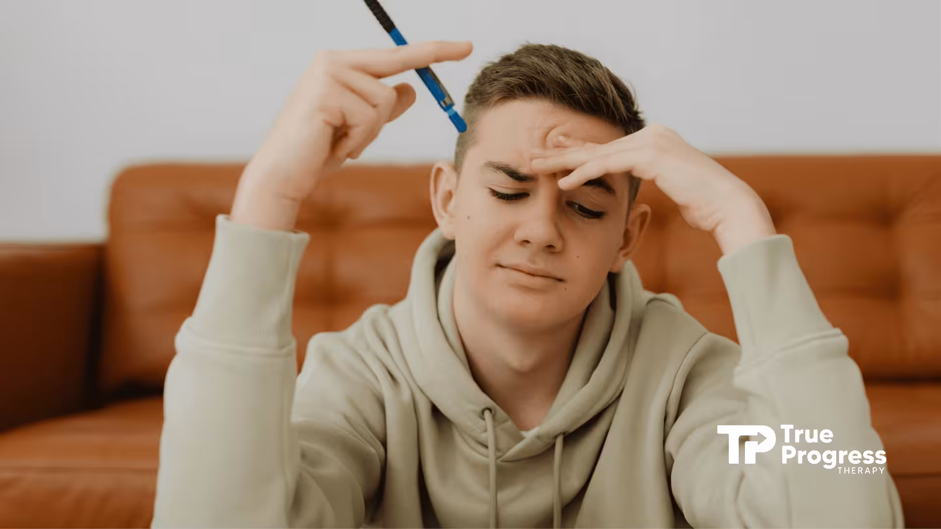 A frustrated RBT student in a hoodie holding a pen while studying on a couch at home in New Jersey.