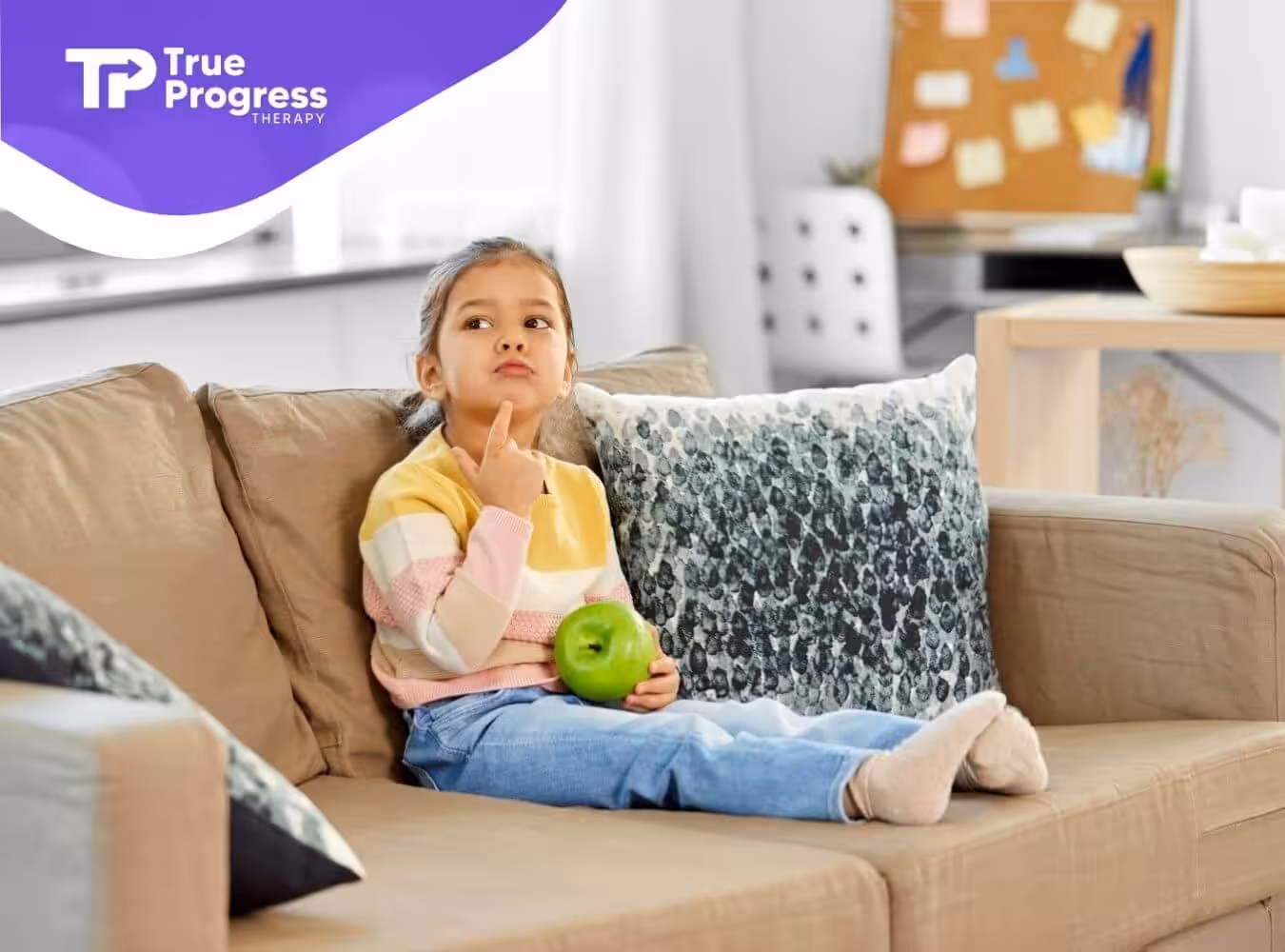 A girl with autism sitting on a couch holding a green apple and thinking with her finger on her chin.