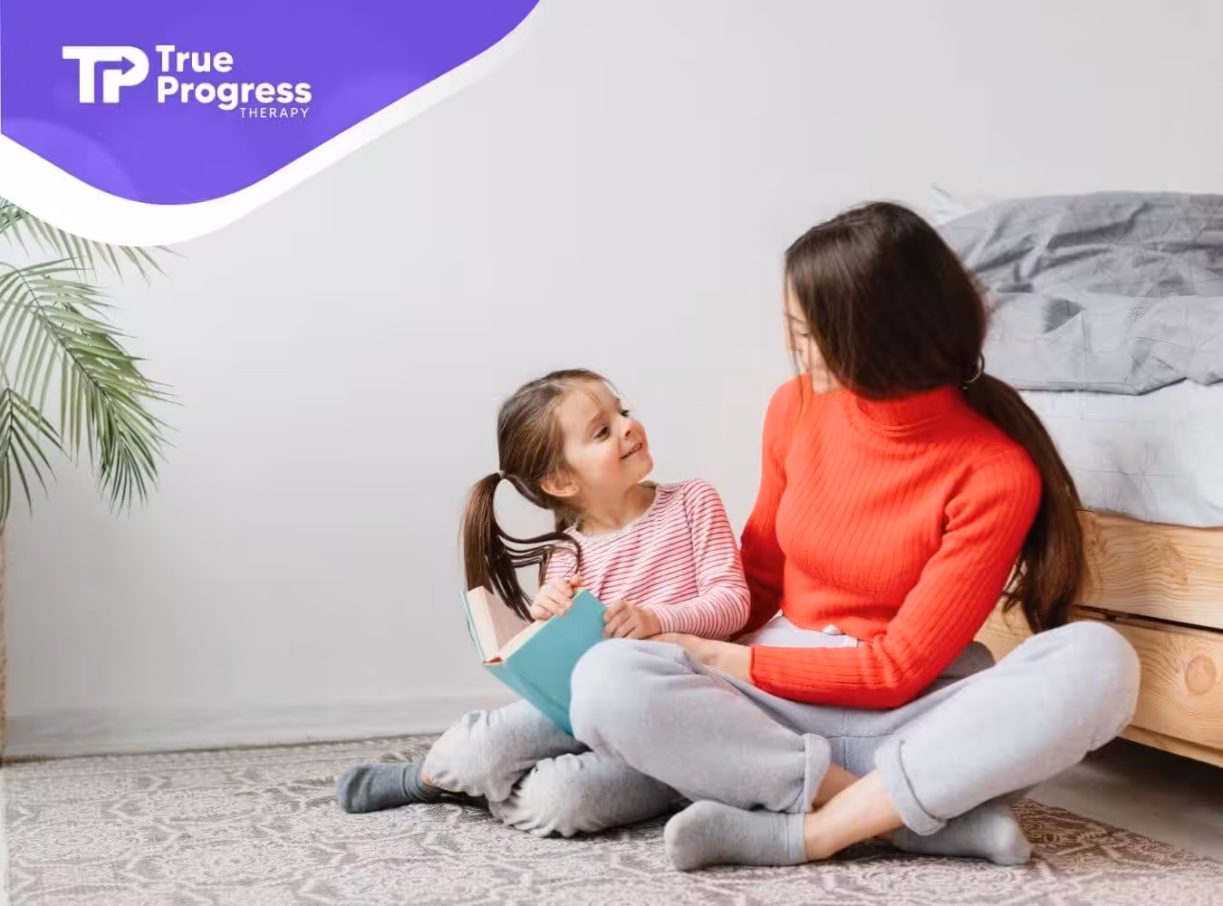 A BCBA with autistic girl smile at each other while sitting on the floor with a book.