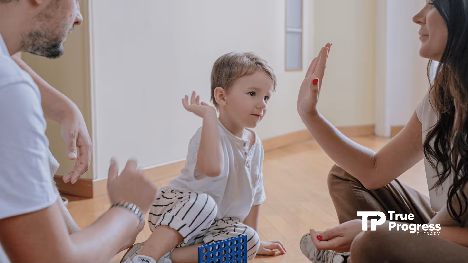 A child in striped pants and white shirt raises a hand for a high-five, smiling with two adults on a wooden floor.