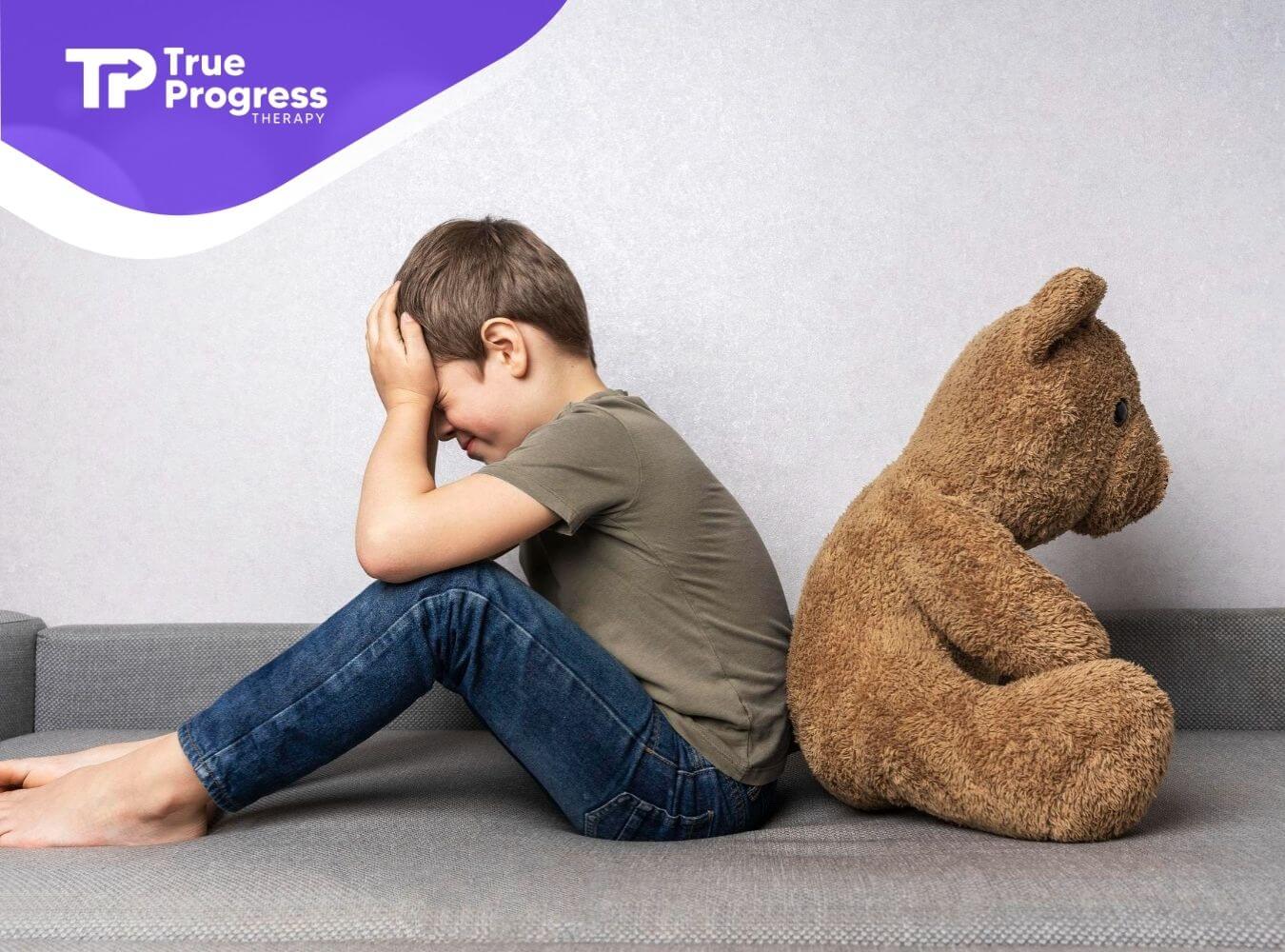 Autistic child sitting with head in hands beside a teddy bear.