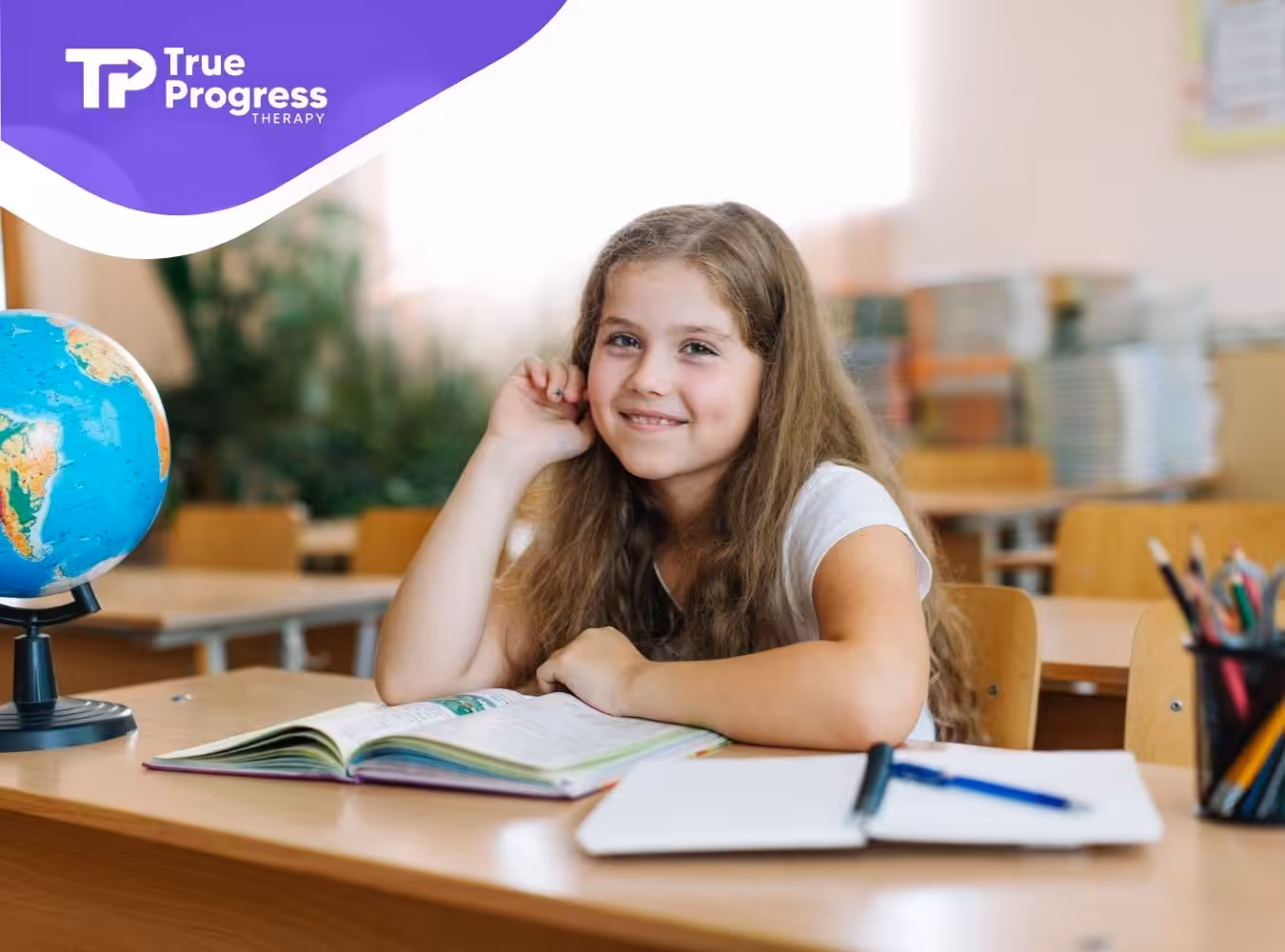 A smiling young girl with long curly hair sits at her desk in a classroom.