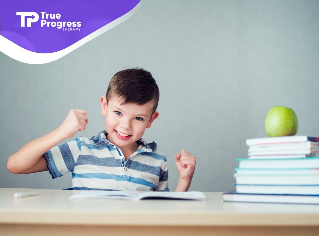 Young child with autism smiling and raising fists while sitting at a desk with books.