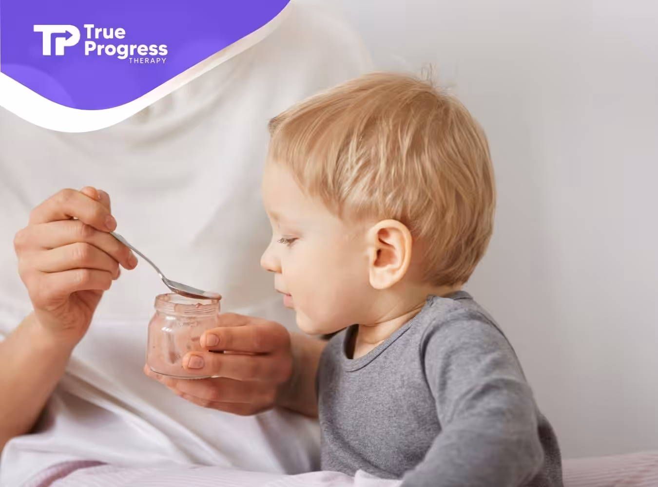 A mother feeding her young child with a spoon from a jar, both smiling.