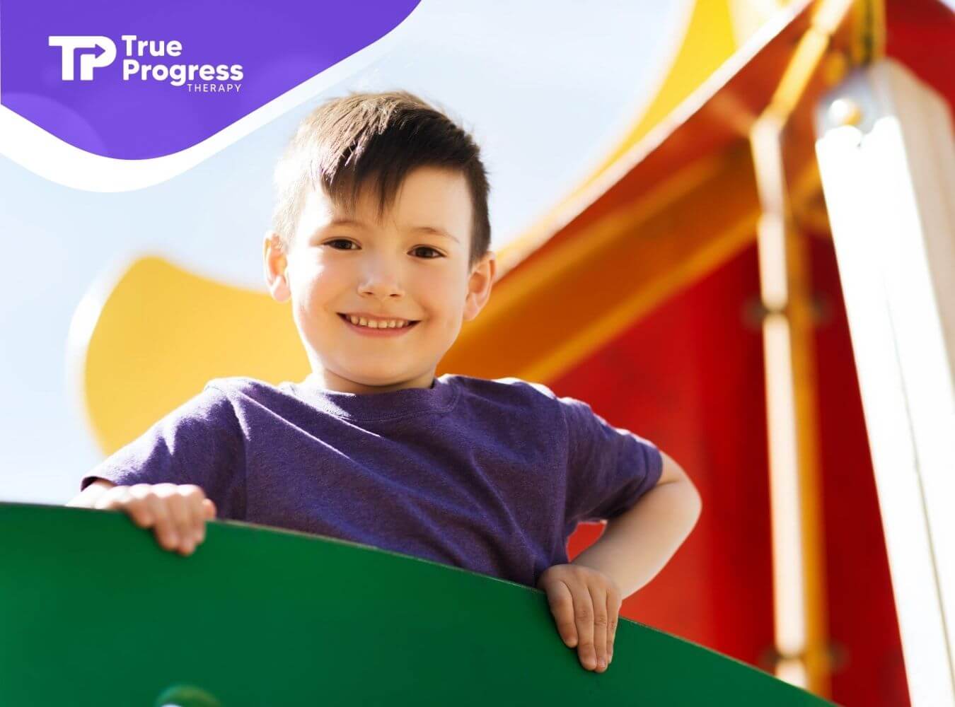 A smiling boy with autism standing on playground equipment with a slide behind him.