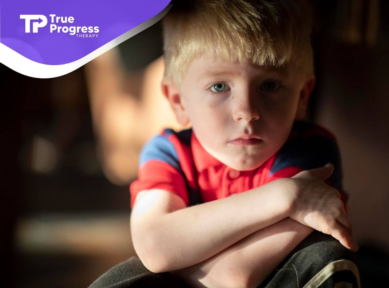 Young child with autism sitting indoors with arms crossed, looking serious.