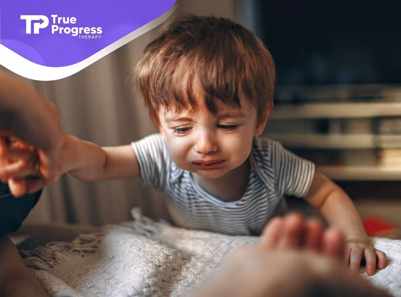 Crying child leans forward on white surface, reaching out to grasp an adult’s hand for comfort and support.
