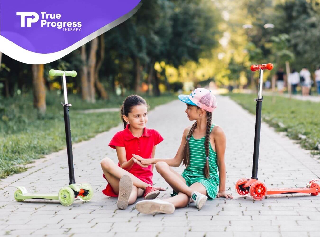 Two autistic girls sitting on a park path beside scooters, holding hands and sharing a moment of friendship and trust.