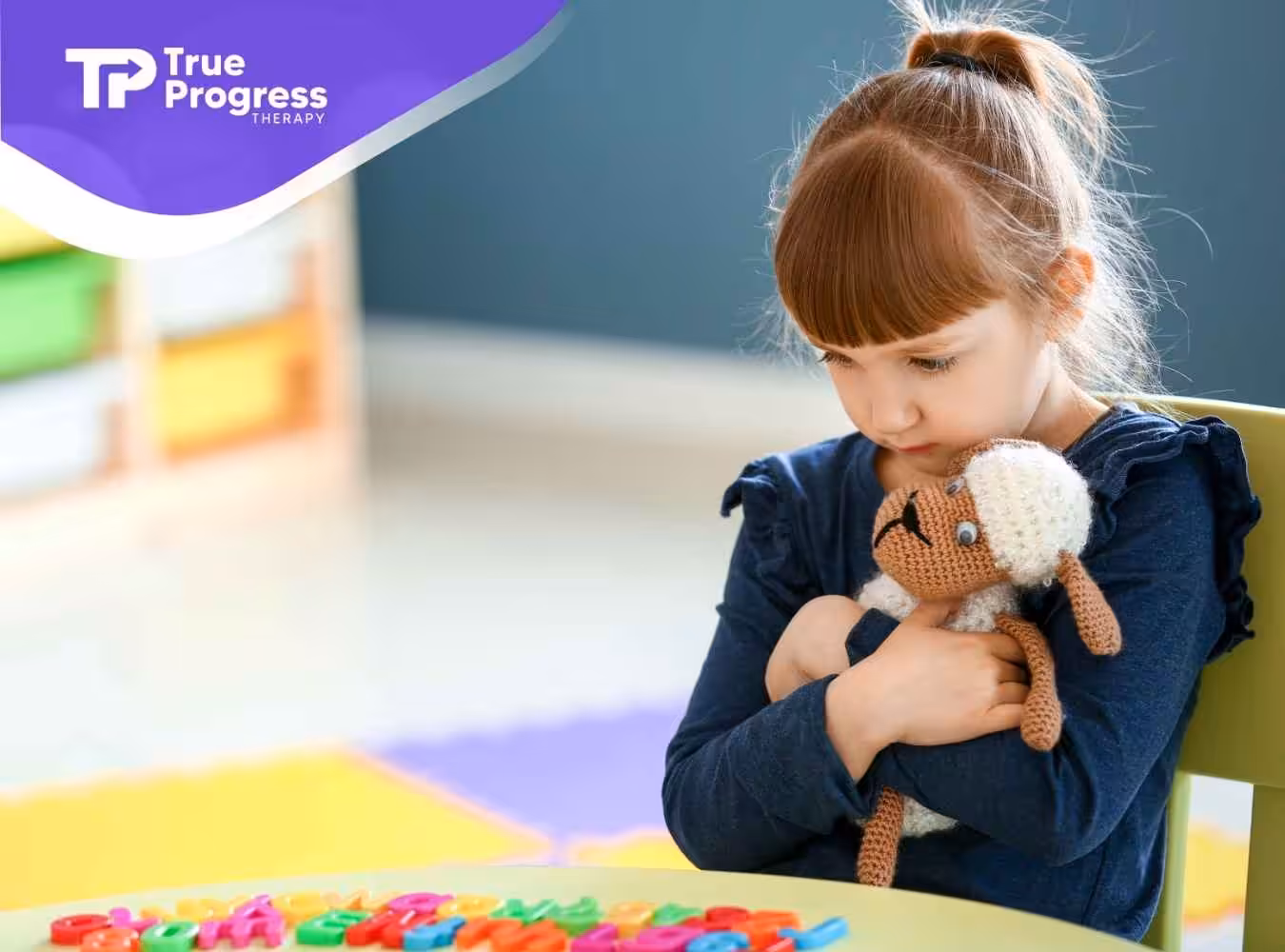 A young girl sits in a classroom, hugging a crocheted lamb toy, appearing thoughtful and calm. Shelves with colorful bins are blurred in the background.