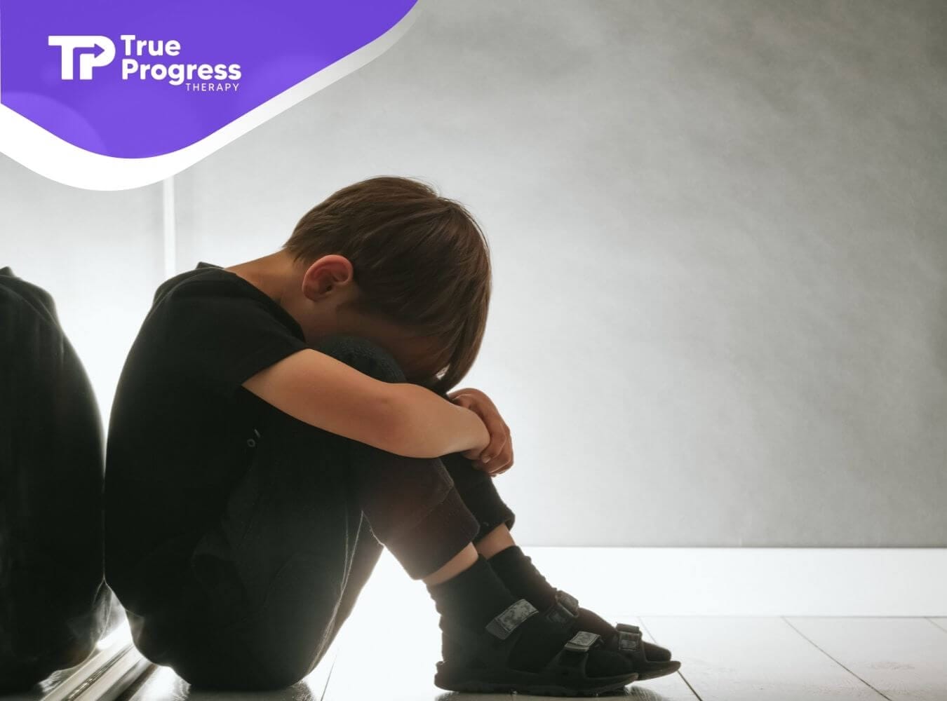 A young boy with autism sitting on the floor with his head down, appearing upset.