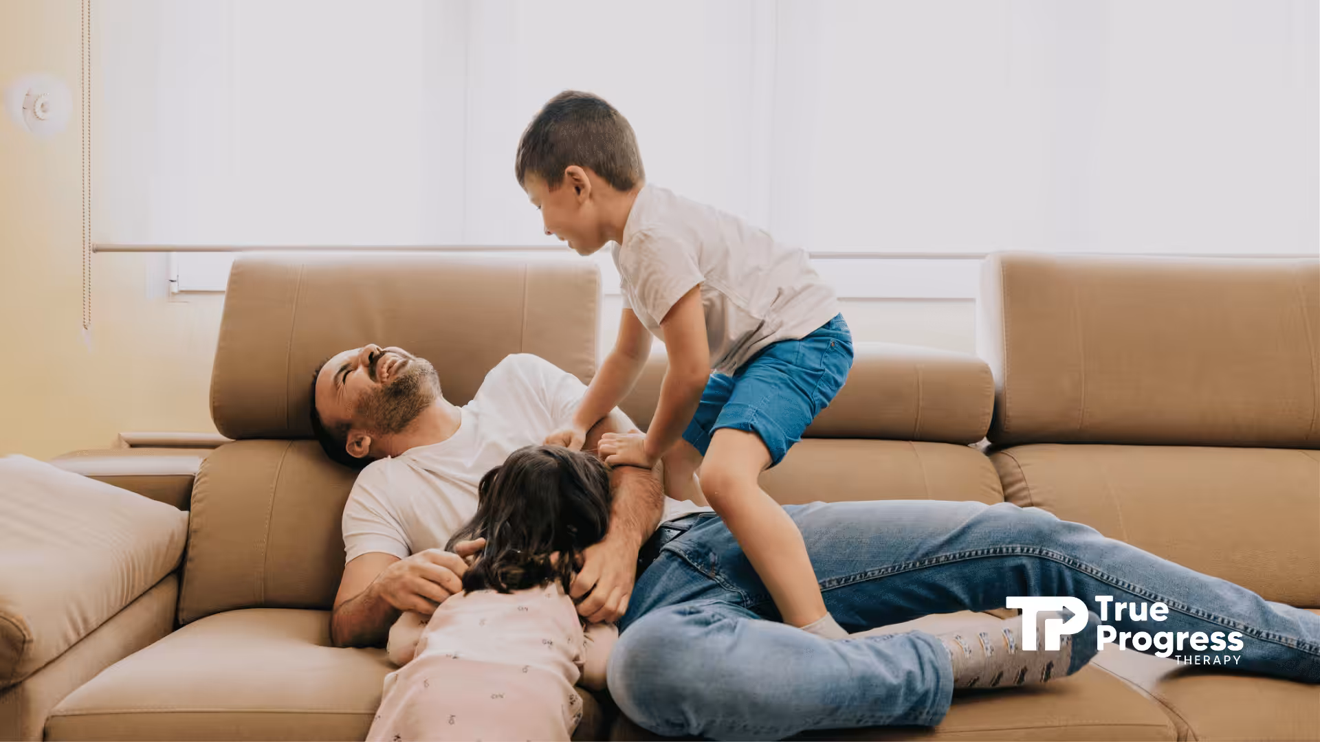 A joyful scene of a father playfully lying on a couch, laughing with his two children. The atmosphere is warm and happy, conveying family bonding.