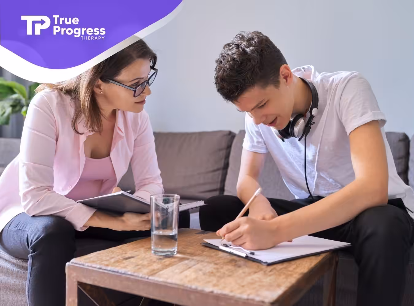 A teenage boy, wearing a noise-cancelling headphones, writing on a paper with his therapist.