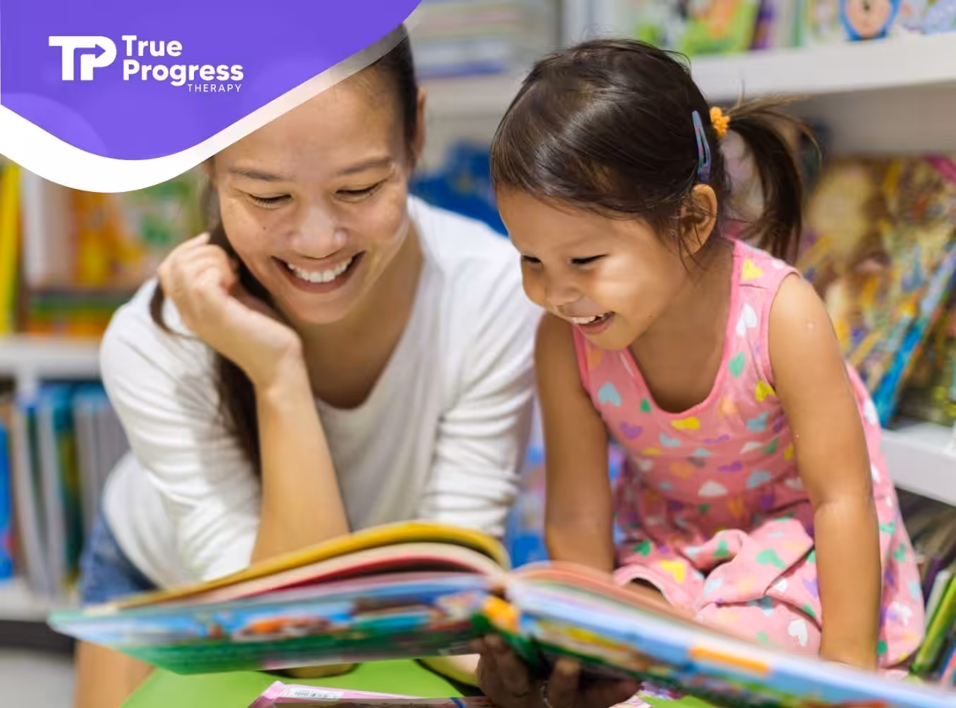 A smiling woman and a young girl read a colorful book together in a bright library. The atmosphere is joyful and educational, with books in the background.