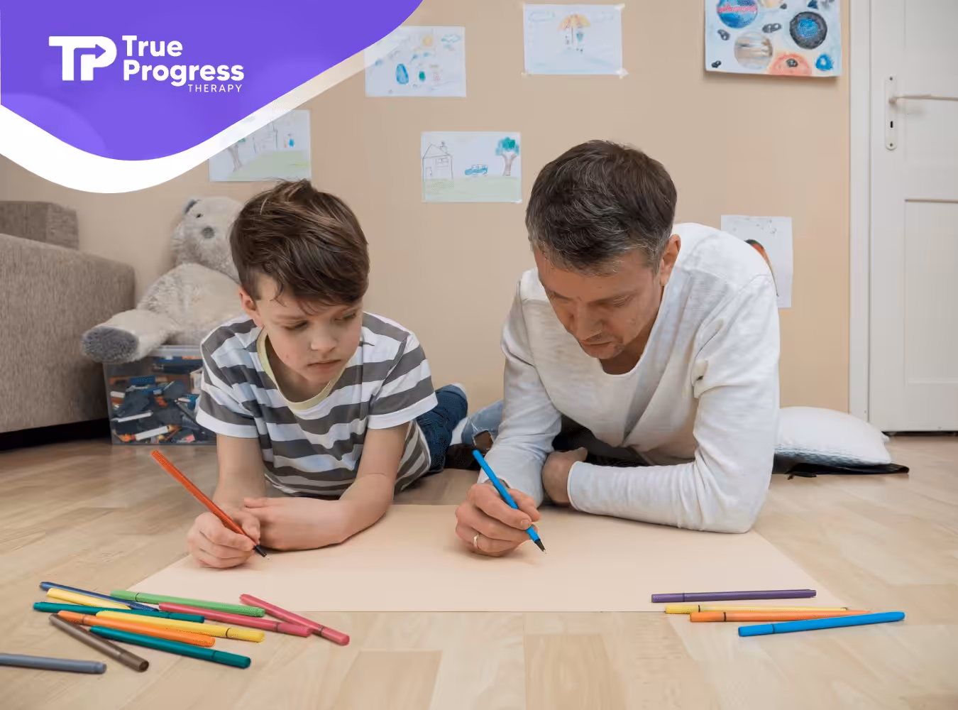 A boy and his therapist are laying on the floor while drawing using colored pencils