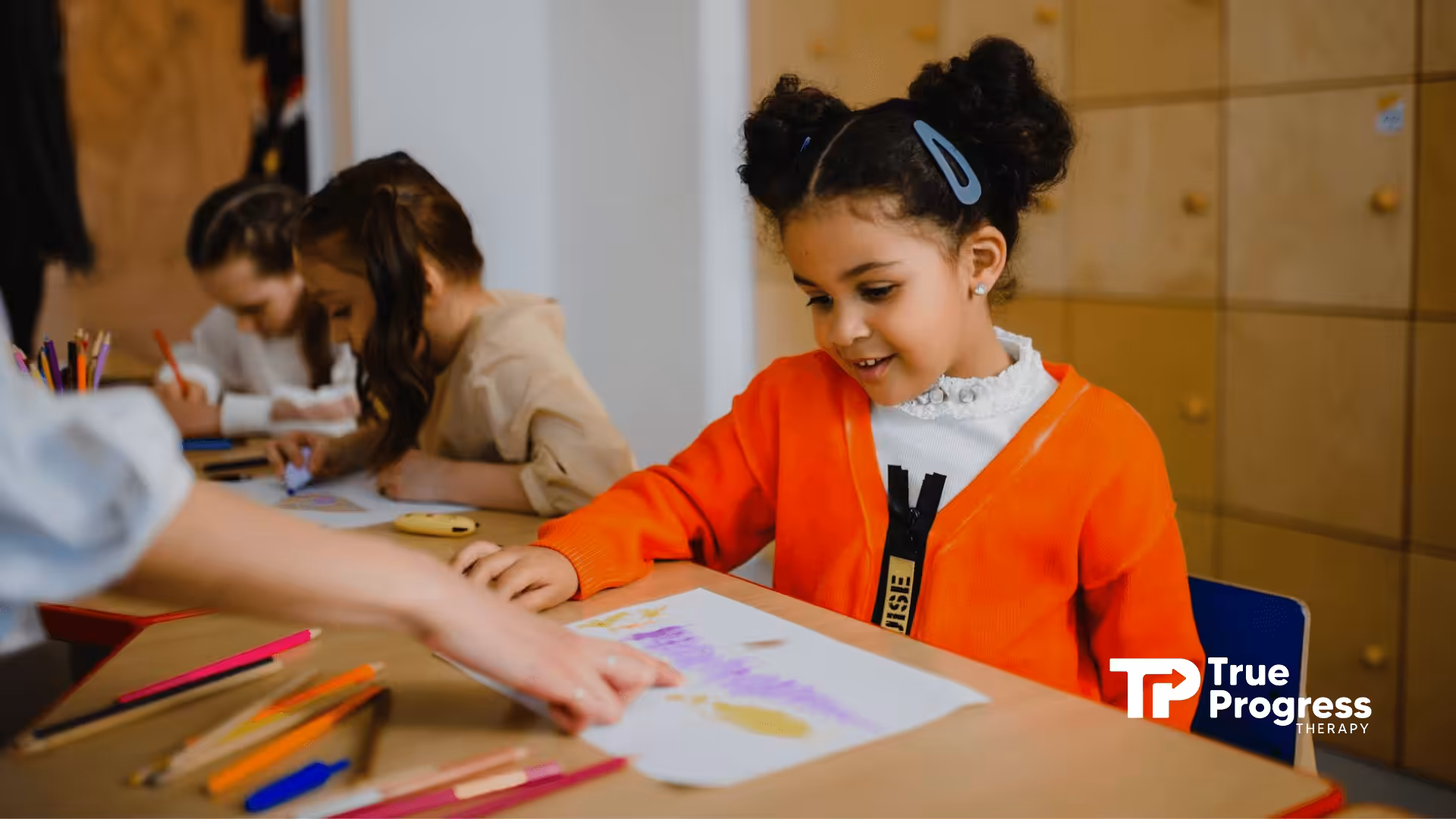 An autistic girl in orange cardigan smiling while drawing at a classroom table during ABA therapy session in NJ.