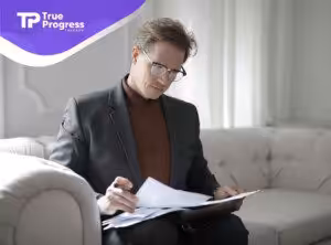 A professional BCBA in glasses reading documents on a sofa, preparing ABA treatment plans for autistic patient in NJ.