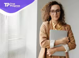 A BCBA in glasses holding a laptop while standing near window blinds as she prepares for a job interview in New Jersey.