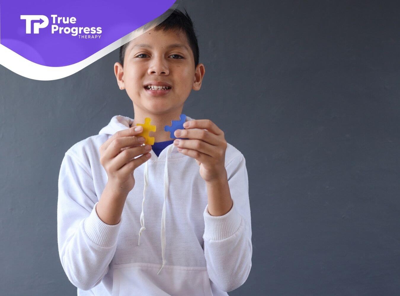 Smiling autistic boy holding two colorful puzzle pieces in front of a gray background, symbolizing learning and connection.