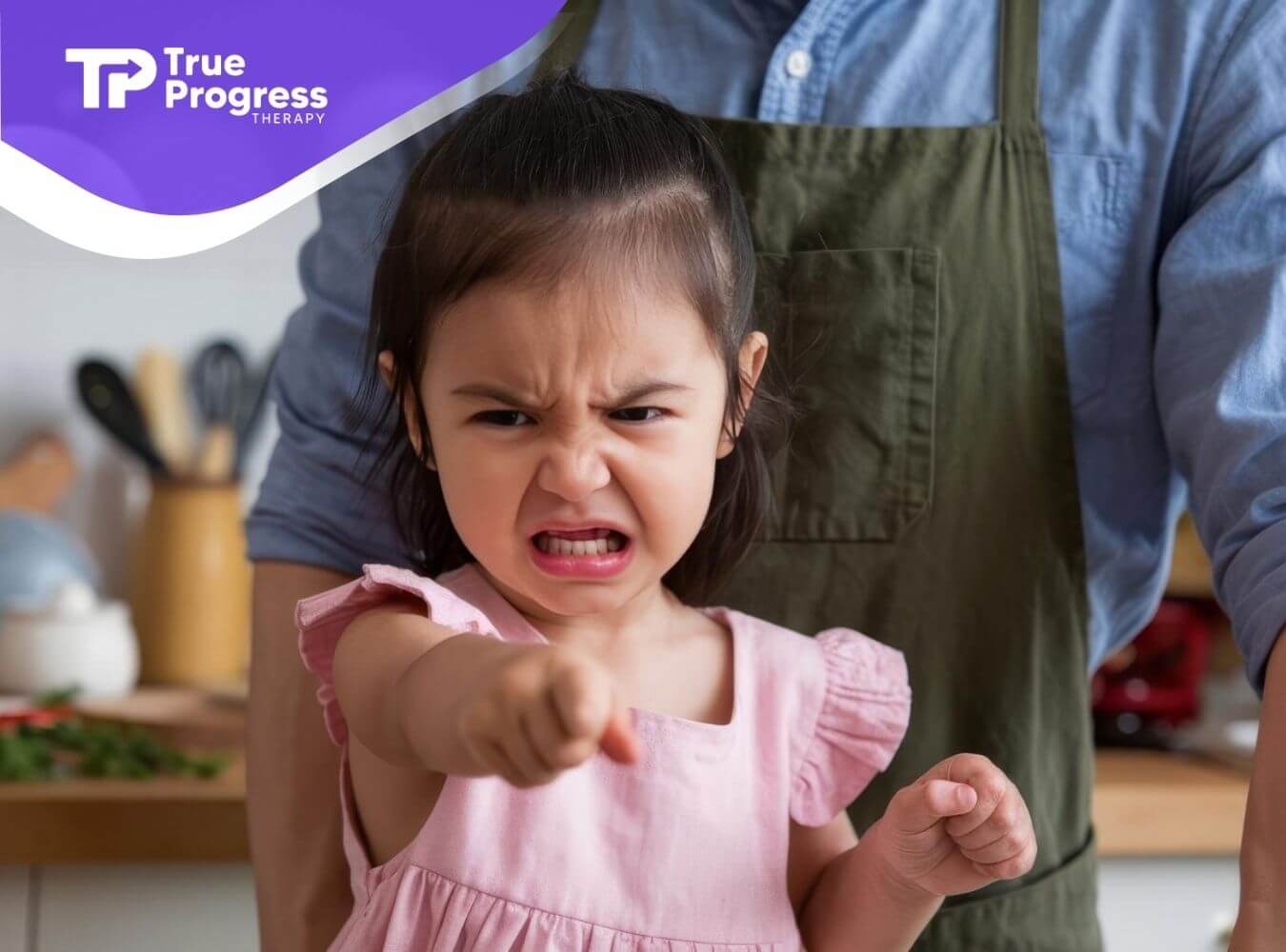 Toddler with autism pointing angrily while a BCBA stands behind in a kitchen.